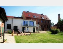 Ein ländliches Anwesen mit einem modernen weißen Anbau und einem größeren roten Backsteingebäude mit Satteldach. Im Vordergrund befindet sich ein gepflegter Garten mit Rasen und gepflasterten Wegen, im Hintergrund eine Spiraltreppe und vereinzelte Blumentöpfe.