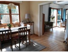 Esszimmer in einem hellen, gemütlichen Haus mit Holzböden; ein Holztisch mit zwei Stühlen und einer Bank steht vor einem großen Fenster mit Blick auf Bäume. Im angrenzenden Raum sind ein Sofa, weitere Sitzmöbel und ein Zugang zu einem sonnigen Bereich mit weiteren Fenstern erkennbar.