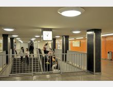 U-Bahn-Station mit einem Treppenaufgang und einer Rolltreppe, umgeben von orangefarbenen Wandfliesen. Eine Uhr und mehrere hell erleuchtete Deckenleuchten befinden sich im Raum, während Personen die Treppen und Rolltreppen benutzen.