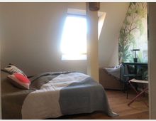 Schlafzimmer unter einer Dachschräge mit einem Einzelbett, dekoriert mit bunten Kissen und einer gemusterten Decke. Ein Dachfenster bringt Tageslicht herein, daneben steht ein kleiner Schreibtisch mit Lampe, und eine Wand ist mit botanischer Tapete verziert.