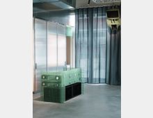 Ein moderner Innenraum mit einem Tresen aus grünem, gitterförmigem Material. Im Hintergrund sind bodenlange, graue Vorhänge sowie eine Metallwand mit lichtdurchlässigen Elementen zu sehen, und oben hängt ein Lautsprecher an der Decke.