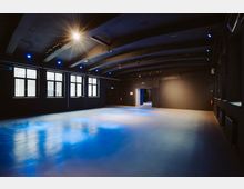 Großer, dunkler Innenraum mit schwarzer Decke und gebogenen Balken, großen Fenstern an der linken Wand und einem reflektierenden, glatten Boden, der in bläulichem Licht schimmert. Im Hintergrund führt eine Tür in einen weiteren Raum.