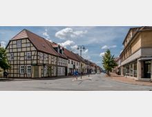 Straßenszene in einer deutschen Kleinstadt mit einem Fachwerkhaus auf der linken Seite, modernen Gebäuden auf der rechten Seite und einer schmalen Verkehrsinsel mit Straßenschildern in der Mitte. Der Himmel ist leicht bewölkt, und einige Autos sowie ein Fahrradfahrer sind auf der Straße zu sehen.