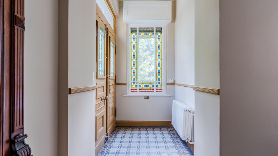 Schmaler Flur im Altbau mit hellem Holz-Eingangstür, hohen weißen Wänden mit Holzleisten und dekorativem Fliesenboden in Blau-Weiß. Am Ende befindet sich ein großes Buntglasfenster mit geometrischem Muster, daneben ein weißer Heizkörper.