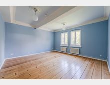 Leerer Raum mit hellblauen Wänden, breiten Holzdielen und hoher Decke mit Stuckleisten und zwei runden Deckenleuchten. Zwei hohe Fenster an der Stirnwand lassen Tageslicht herein; darunter stehen weiße Heizkörper.