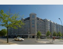 Mehrstöckiges, modernes Wohn- und Geschäftshaus in grauer Fassade mit vielen Fenstern und kleinen Giebeln, gelegen an einer Straßenecke mit Gehwegen und parkenden Autos. Im Vordergrund ein einzelner Baum, ein vorbeifahrendes Taxi und Fußgänger.