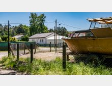 Ein ländliches Grundstück mit einem weißen Einfamilienhaus im Hintergrund, umgeben von Zäunen und Bäumen. Im Vordergrund steht ein gelbes Boot auf einem Anhänger hinter einem Maschendrahtzaun.