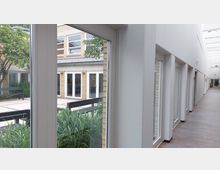 Blick durch eine Glasfront auf einen Innenhof mit Pflanzbeeten, Sitzbank und Gebäudefassade aus hellen Ziegelsteinen mit weißen Türen. Rechts verläuft ein moderner, lichtdurchfluteter Korridor mit Holzboden und weißen Wänden.