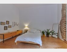Minimalistisch eingerichtetes Schlafzimmer mit einem Holzbett und weißer Bettwäsche, einem hölzernen Lowboard an der linken Wand, dekorativen Pflanzen in Töpfen, einem großen Standspiegel rechts und einem teilweise freigelegten Mauerstück. Der Boden besteht aus Holz, und an der Wand hängen gerahmte Bilder und kleine Dekorationen.