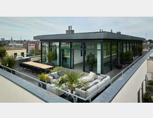 Moderne Dachterrasse mit einem verglasten Gebäude, das große bodentiefe Fenster hat. Vor dem Gebäude befinden sich eine Sitzgruppe mit grauen Sofas, ein Holztisch mit Stühlen sowie Pflanzkübel mit Bambus und Palmen. Im Hintergrund ist eine urbane Stadtlandschaft sichtbar.