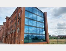 Eine ehemalige Backsteinfabrik mit einem modernen Anbau aus Glasfassaden, die den Himmel und umliegende Gebäude spiegeln. Im Hintergrund sind Wiesen und weitere Industriegebäude zu sehen.