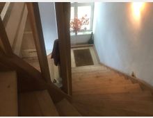 Holztreppe in einem Innenraum mit einer weißen Wand und sanfter Beleuchtung. Am unteren Ende der Treppe befinden sich ein Teppich, ein Fenster mit einem Baumzweig in einer Vase und eine geöffnete Tür.