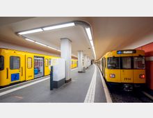 U-Bahn-Station mit zwei gelben U-Bahn-Zügen; einer steht rechts mit der Anzeige „U7 Rudow“, während der andere links geöffnet ist. Die Station hat glatte, weiße Decken, Säulen und graue Böden mit markierten Bahnsteigkanten.
