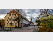 Straßenszene mit einem gelben Haus auf der linken Seite, einer Reihe bunter Gebäude mit Schaufenstern, sowie einem Kirchturm mit spitzer Spitze im Hintergrund. Blendende Herbstbäume und Straßenlaternen mit Blumen dekorieren den Gehweg.