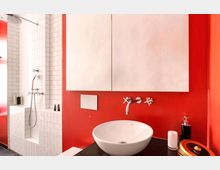 Modernes Badezimmer mit auffälliger roter Wandgestaltung, einem runden weißen Waschbecken auf einer dunklen Arbeitsplatte und einer Wandarmatur mit Kreuzgriffen. Im Hintergrund eine Dusche mit weißen Fliesen und verchromter Armatur.