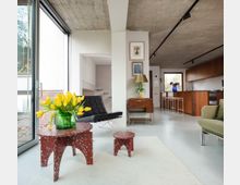 Open-plan modern living room and kitchen with a raw concrete ceiling, large floor-to-ceiling sliding glass doors, and white floors. The space includes a black lounge chair, green sofa, terrazzo-style red tables on a light rug, and a wood-fronted kitchen with bar stools along the counter.