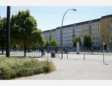 Straßenszene mit mehrstöckigen Wohnhäusern in Gelb- und Weißtönen im Hintergrund, einer Baumreihe entlang der Straße, einer Bushaltestelle und einem Radfahrer im Vordergrund. Links befindet sich eine bepflanzte Grünfläche mit Blumen und Sträuchern.