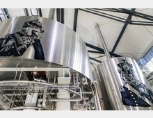 Großer industrieller Braukessel aus Edelstahl in einer modernen Brauerei, verziert mit künstlerischen Darstellungen eines Wolfes und eines Tigers in Uniform. Im Hintergrund sind Dachträger und Rohrleitungen sichtbar.