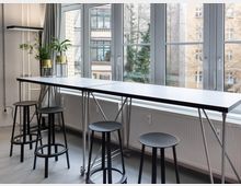 Heller Innenraum mit langem, schmalem Stehtisch auf Metallgestell und mehreren schwarzen Hockern entlang einer Fensterfront. Große, mehrteilige Fenster mit Blick auf benachbarte Gebäude; auf der Tischplatte stehen Pflanzen in Töpfen.