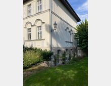 Seitenansicht eines hellen, historischen Gebäudes mit hohen Fenstern und dekorativen Fassadenelementen. Der Sockelbereich besteht aus Naturstein, umgeben von gepflegtem Grün und Büschen in einem Garten.