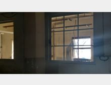 Fenster in einem verlassenen, industriellen Gebäude, mit einer einfachen Metallrahmung und Blick auf eine weitere Wand mit Rohren und einem teilweise verhängten Fenster. Der Raum wirkt durch den Lichteinfall aus den Fenstern hell, aber verlassen.