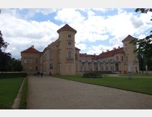 Schlossanlage mit hellgelbem Gebäude im Barockstil, gekennzeichnet durch Türme mit roten Dächern, Säulengängen und symmetrischer Fassade. Im Vordergrund ein gepflegter Garten mit Rasenflächen, Wegen und Statuen.