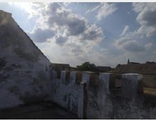 Eine verfallene Mauer mit Zinnen und abgeblättertem Putz im Vordergrund, daneben das Dach eines angrenzenden Gebäudes. Im Hintergrund sind Dächer eines Dorfes, Bäume und ein Kirchturm unter einem teils bewölkten Himmel zu sehen.