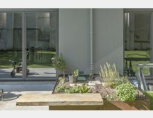 Moderne Terrasse mit Hochbeet im Vordergrund, bepflanzt mit Kräutern und kleinen Pflanzen. Im Hintergrund eine Glas-Schiebetür, die den Blick auf einen Rasen und ein Kinderdreirad freigibt.