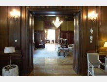 Raum in einem historischen Gebäude mit Holzwänden, reich verzierten Türrahmen und einem Kronleuchter in der Mitte. Im Hintergrund sind ein weiterer Raum mit Polstermöbeln, Teppich und dekorativen Tischlampen sichtbar.