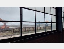 Blick durch ein großes Industriefenster mit Metallrahmen auf ein Außengelände mit Bahnstrecken, rostenden Gebäudestrukturen, einem blauen Kran und einer grauen, bewölkten Landschaft im Hintergrund.