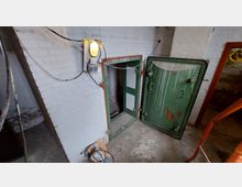 Industrie- oder Kellerraum mit weiß gestrichenen Ziegelwänden, einer geöffneten grünen Metalltür und freiliegenden Kabeln. Eine Wandlampe beleuchtet den Bereich, neben der Tür führt ein schmaler Zugang in einen weiteren Raum.