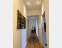 Heller Flur in einer Wohnung mit Holzboden und weißen Wänden, an denen ein buntes Gemälde hängt. Im Hintergrund steht ein Holzmöbelstück mit Dekorationen, seitlich sind geöffnete Türen sichtbar.