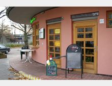 Straßenszene mit einem kleinen Café mit rosafarbener Fassade und Holztüren, vor dem Eingang stehen ein Klapptisch, Plastikstühle und eine Kreidetafel mit Angeboten. Im Hintergrund sind eine U-Bahn-Station sowie parkende Autos sichtbar.