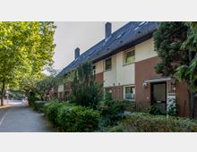 Reihenhaus mit braunen und weißen Fassaden, schrägem Dach und mehreren Fenstern, umgeben von dichtem Grün mit Bäumen und Büschen entlang eines Gehwegs. Im Hintergrund ist eine Straße mit leichter Verkehrsabsperrung erkennbar.