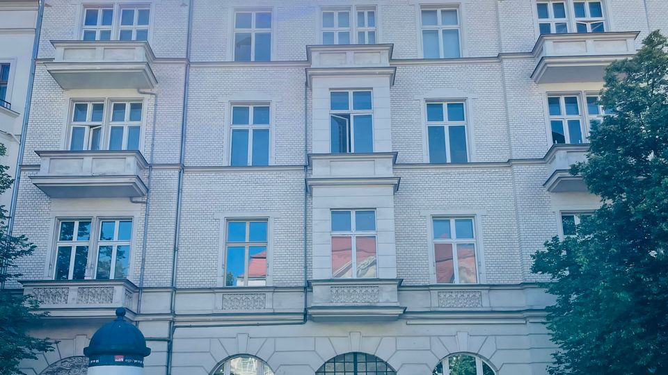 Mehrstöckiges helles Backsteingebäude mit symmetrischer Fassade, hohen Fenstern, kleinen Balkonen und zwei spitzen Giebeln; im Erdgeschoss befindet sich ein zentraler Eingangsbereich mit Bogenfenster. Davor verläuft eine Straße mit einem Litfaßsäulen-Plakat und Bäumen am rechten Rand unter blauem Himmel mit Sonne.