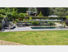 Private Gartenanlage mit modernem Swimmingpool, umgeben von Steinmauern und reichlich bepflanzten Beeten. Im Hintergrund sind ein modernes Holzhaus sowie Bäume und Sträucher zu sehen, während im Vordergrund Kinder auf der Terrasse spielen.