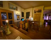 Gemütlicher Restaurantinnenraum mit Holztischen und -stühlen, einer Theke mit Barhockern sowie warmem Kerzen- und Lampenlicht. An den hellen Wänden hängen Bilder und Tafeln mit Speisekarte, daneben eine Holztür und ein Durchgang in einen weiteren Gastraum.