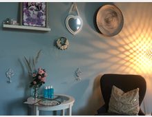 Ein kleiner, gemütlicher Innenraum mit pastellblauer Wand, dekoriert mit einem Herzspiegel, einem modernen Kunstbild und weiteren Wanddekorationen. Vor der Wand stehen ein weißer Tisch mit Blumen und blauen Flaschen sowie ein schwarzer Sessel mit einem beigen Kissen. Eine Lampe wirft ein warmes, hölzernes Lichtmuster auf die Wand.