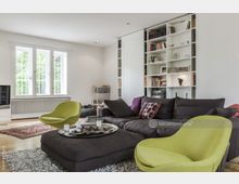 Modernes Wohnzimmer mit großer grauer Eckcouch, zwei hellgrünen Polstersesseln und einem Teppich auf Holzboden. Im Hintergrund ist ein Bücherregal, große Fenster mit Blick ins Grüne und ein Fernseher zu sehen.