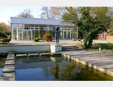 Ein modernes, rechteckiges Gebäude mit Glasfassade, umgeben von Bäumen und einem gepflasterten Außenbereich. Im Vordergrund ein flaches Wasserbecken mit Stegen und eine Skulptur auf einem Sockel.