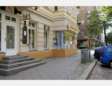 Straßenszene mit gelb-beigem Eckgebäude, das Geschäfte im Erdgeschoss beherbergt, darunter ein Weinladen und ein Geschäft mit der Aufschrift „schöner schlafen“. Im Vordergrund Gehweg und parkende Autos sowie Straßenbäume im Hintergrund.