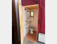 Kleines Badezimmer mit offener Holztür, ausgestattet mit einer Toilette, kleinem Waschbecken und einem hoch an der Wand montierten Spülkasten. Die Wände sind mit weißen Fliesen verkleidet, der Boden besteht aus orangefarbenem Belag, und ein kleines Fenster sorgt für Tageslicht.