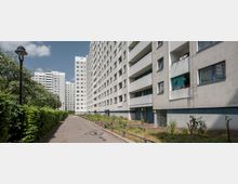 Mehrgeschossige Wohnhäuser in einem urbanen Umfeld, mit breiten Balkonen und zahlreichen Fenstern. Vor den Gebäuden verläuft ein schmaler Gehweg, gesäumt von Hecken und Bäumen; einzelne Straßenlaternen sind sichtbar.