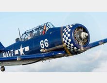 Blaues historisches Propellerflugzeug mit der Aufschrift "NAVY" und der Nummer "66" auf der Seite, versehen mit einem karierten Muster um den Motor, im Flug vor einem wolkigen Himmel. Zwei Personen sind in der verglasten Kabine zu sehen.