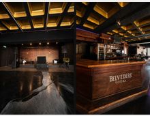 Innenraum eines Musikclubs mit kleiner Bühne, Lautsprechern und Schlagzeug vor gemusterter Rückwand sowie einer Decke mit goldfarbenen Kassetten. Daneben eine lange Bar aus dunklem Holz mit Flaschenregalen und dem Schriftzug „Belvedere Vodka“ an der Front.