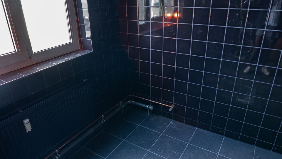 Ecke eines innenliegenden Raums mit dunkelblauen, glänzenden Wandfliesen im Raster und passenden Bodenfliesen. Links ein Fenster mit breiter Fensterbank, darunter ein Heizkörper sowie sichtbare Rohrleitungen entlang der Wand.