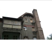 Ein altes, mehrstöckiges Gebäude mit grauem Putz und Backsteinelementen, teils zugemauerten Fenstern und einem hohen Schornstein aus Backstein an der rechten Seite. Die Fassade zeigt verblasste und beschädigte Details, einschließlich eines großen, geschlossenen Eingangsbereichs.