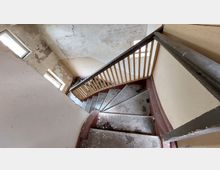 Treppe in einem verlassenen Gebäude mit abblätterndem Putz an den Wänden, einem Geländer aus Holz und einer Metallhandlauf. Der Boden der Treppe und der Bereich drumherum wirken verschmutzt und alt, mit sichtbaren Spuren von Verfall.