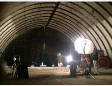 Großer, halbrund gewölbter Innenraum mit sichtbaren Stahlträgern an der Decke, genutzt als Film- oder Fotoset. Im Vordergrund stehen Beleuchtungsequipment und Kameras, während im Hintergrund eine Person beleuchtet ist und ein roter Traktor am rechten Rand sichtbar ist.