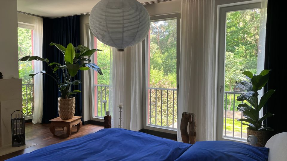 Modernes Schlafzimmer mit großem Bett und blauer Bettwäsche, einer runden Papier-Hängeleuchte sowie Holzboden. Bodentiefe Fenster mit weißen Vorhängen öffnen den Blick auf einen Balkon mit Geländer und viel grünem Baumbestand; mehrere große Zimmerpflanzen stehen am Fenster.